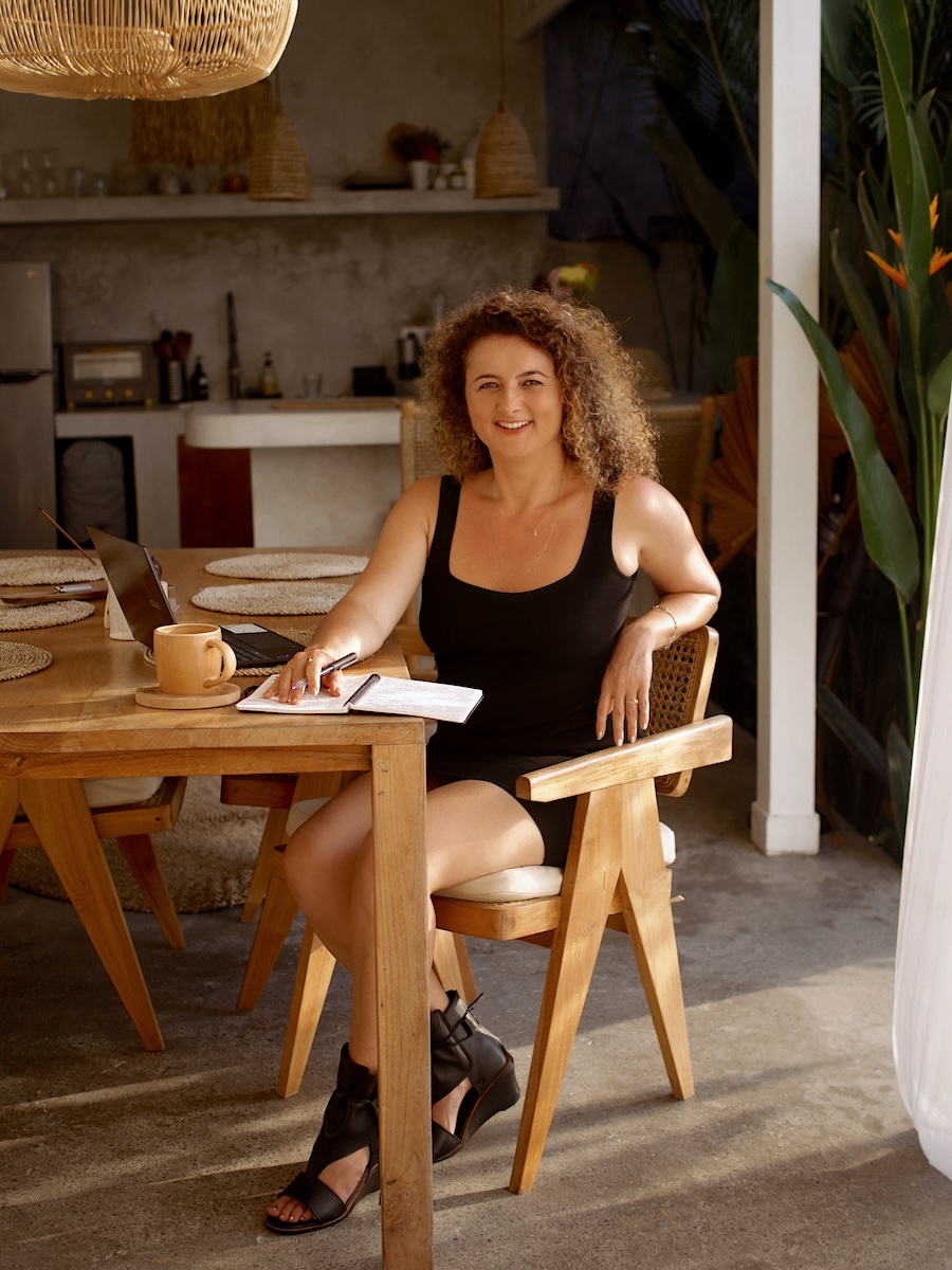 Dijan in a black tank dress seated at a wooden dining table with a laptop and coffee, in soft daylight.