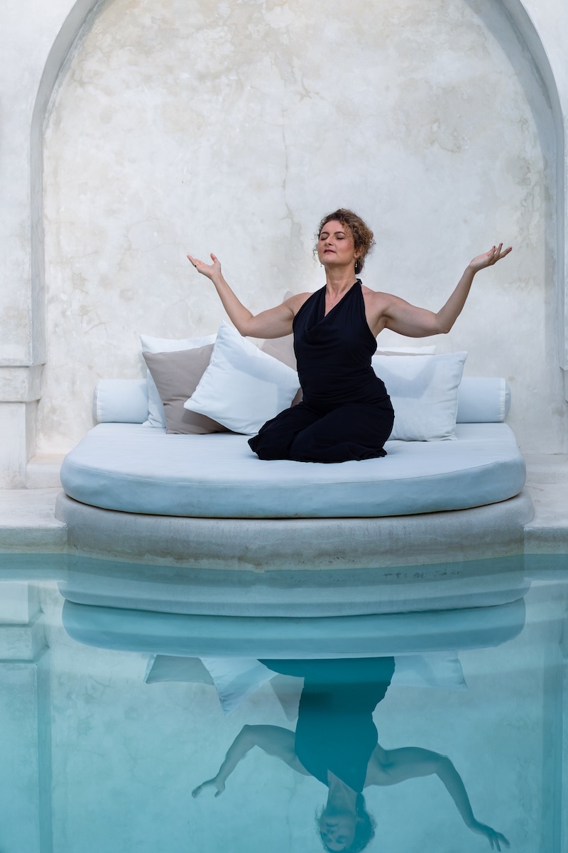Dijan kneeling on a poolside daybed with her arms lifted and palms open, eyes closed, reflected in still water